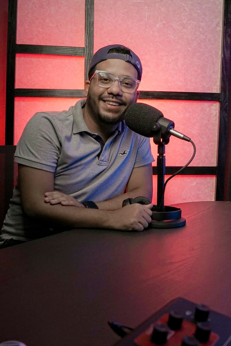 A smiling host sits at a microphone in a studio lit in red.