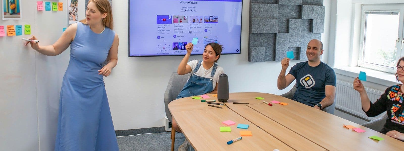 A woman leads a session at a wall of notes while her team, smiling, holds up cards around a conference table.